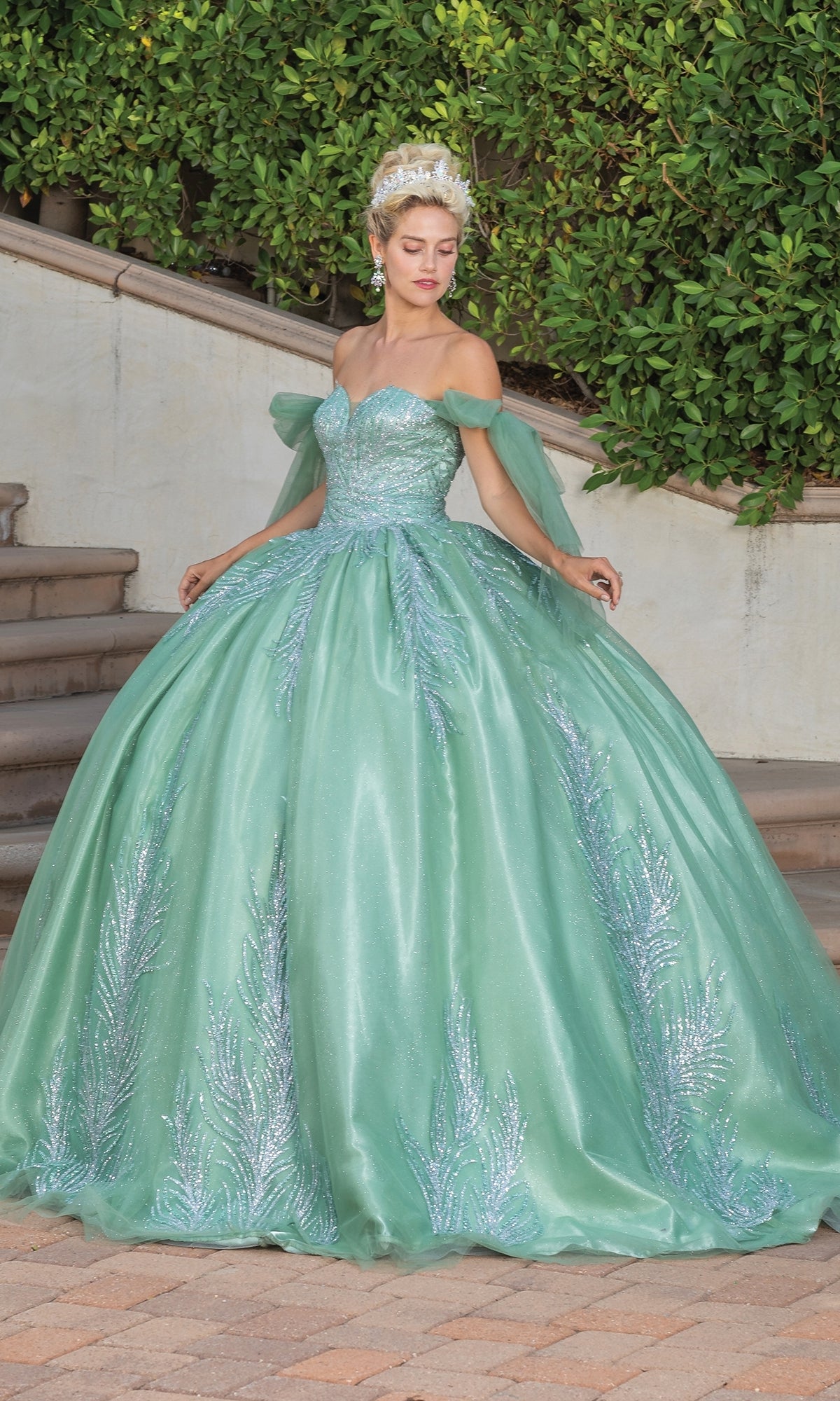 A woman stands outdoors in the Quinceanera Dress 1793 By Dancing Queen, an off-shoulder sage green ball gown with silver embellishments, perfect for special occasions. Greenery and stone steps provide a scenic background.