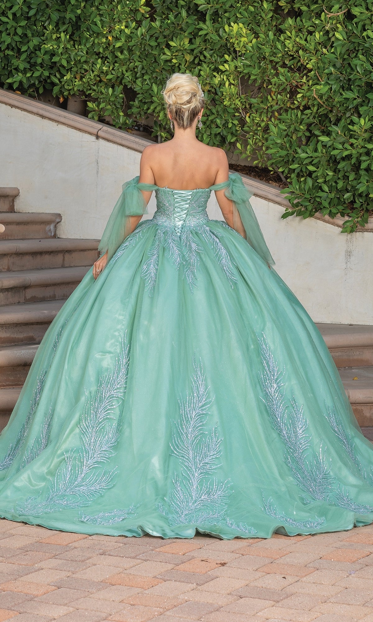 A woman with blonde hair in an updo wears the Quinceanera Dress 1793 By Dancing Queen, an off-shoulder mint green ball gown with silver feather-like patterns, standing on a stone path near steps and greenery.