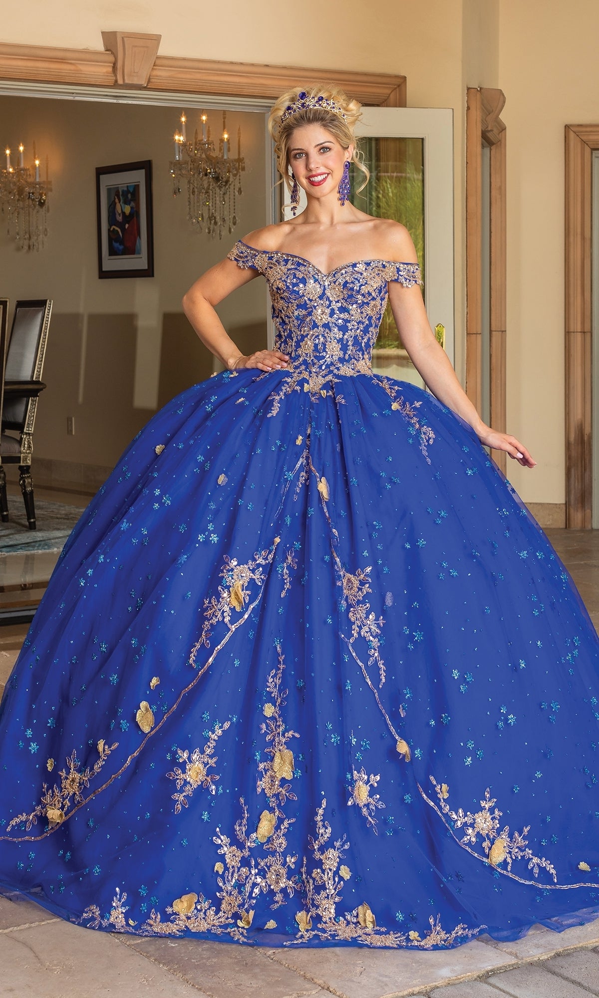 A woman stands indoors by open doors wearing the Quinceanera Dress 1796 By Dancing Queen, a royal blue off-shoulder ball gown featuring gold and silver lace embroidery.