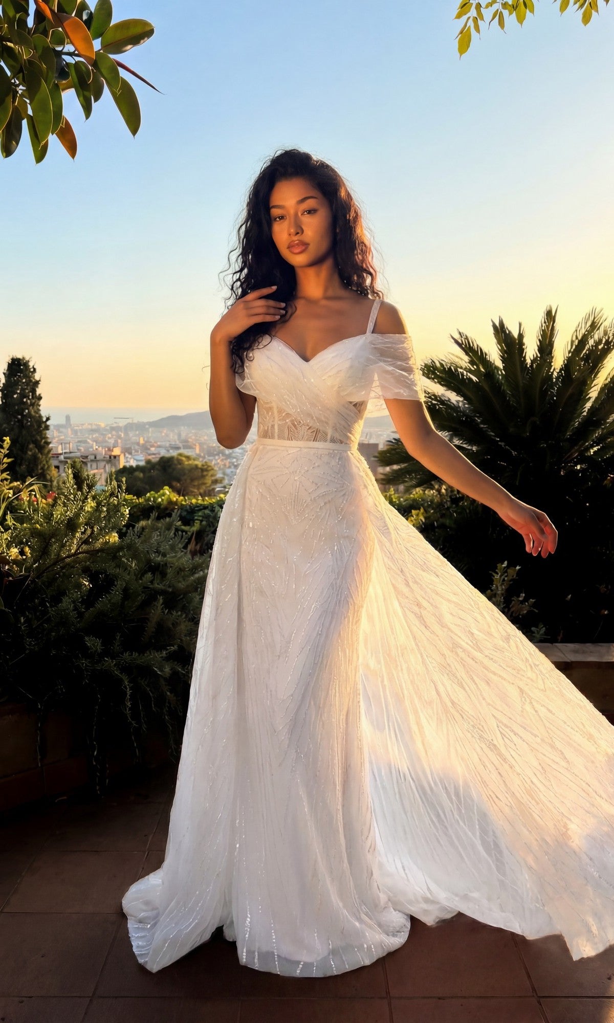 A woman in the off-white Long Formal Dress: Chicas 17990 stands outdoors at sunset, amidst greenery with a cityscape behind her.