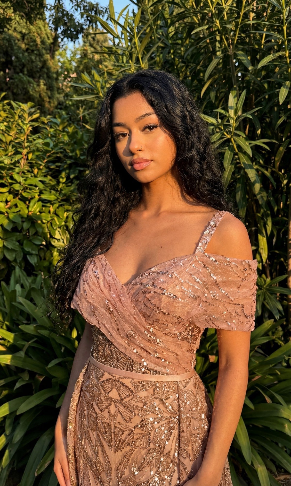 Woman with long dark hair wears the Chicas 17990 Long Formal Dress, a sequined, off-shoulder pink embellished gown, standing outdoors before green foliage and looking at the camera.