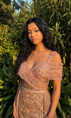 Woman with long dark hair wears the Chicas 17990 Long Formal Dress, a sequined, off-shoulder pink embellished gown, standing outdoors before green foliage and looking at the camera.
