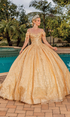 A woman stands on a poolside patio surrounded by greenery, wearing the Quinceanera Dress 1804 By Dancing Queen—a sparkling gold A-line ball gown with a sweetheart neckline—paired with a crown and earrings.