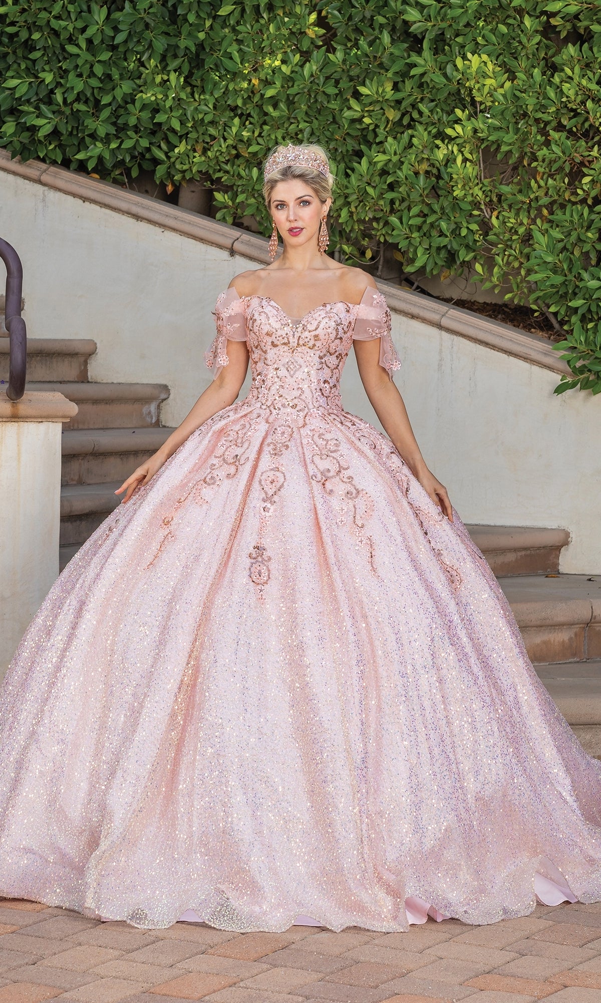 A woman stands outdoors on a stone pathway in the Quinceanera Dress 1804 By Dancing Queen—a pink, sparkly A-line ball gown with a sweetheart neckline and off-the-shoulder sleeves—her tiara completing the elegant look amid greenery and stairs.