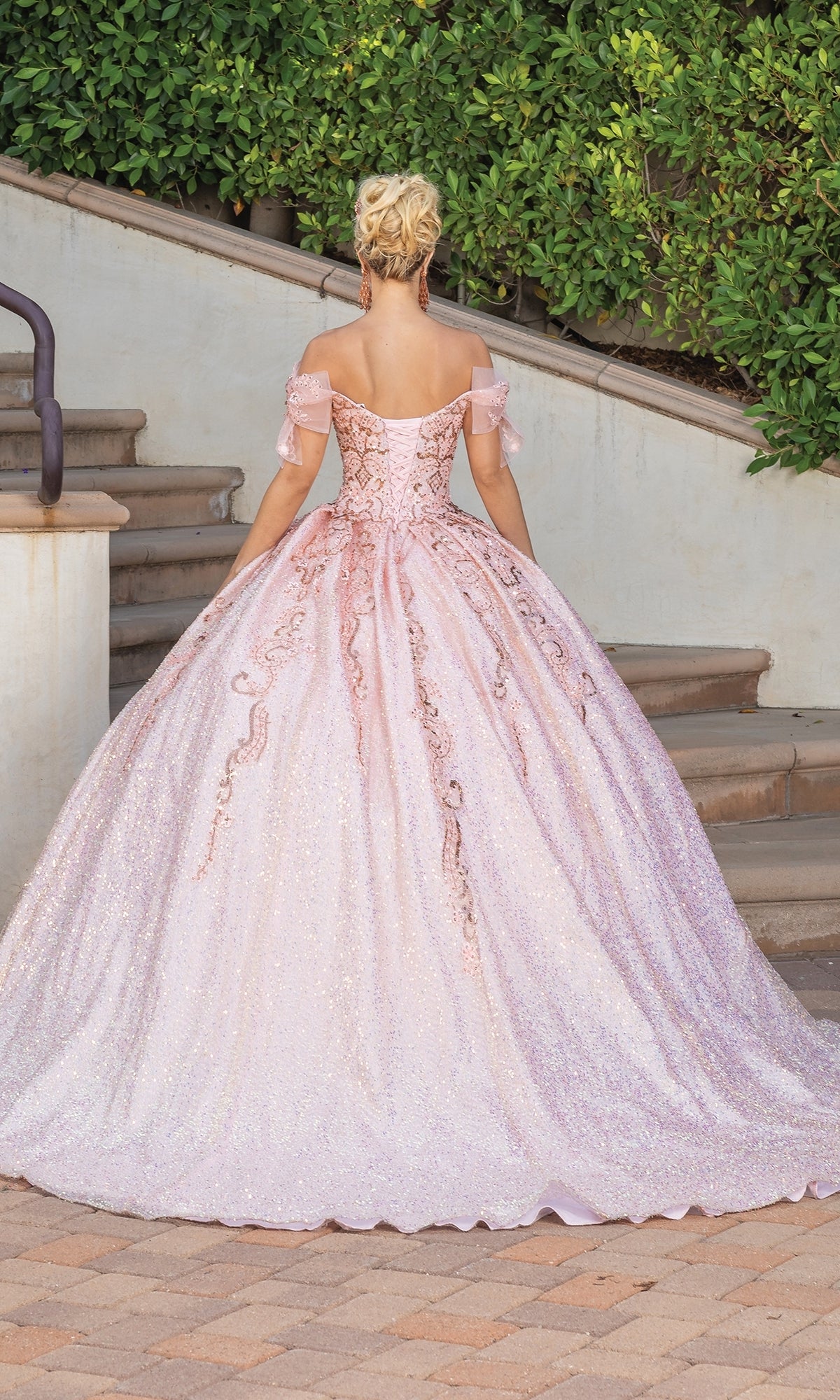 A woman stands outdoors on a stone path, facing away from the camera in front of stairs and greenery, wearing the Quinceanera Dress 1804 By Dancing Queen—a pink, off-the-shoulder, floor-length A-line ball gown with a sweetheart neckline.