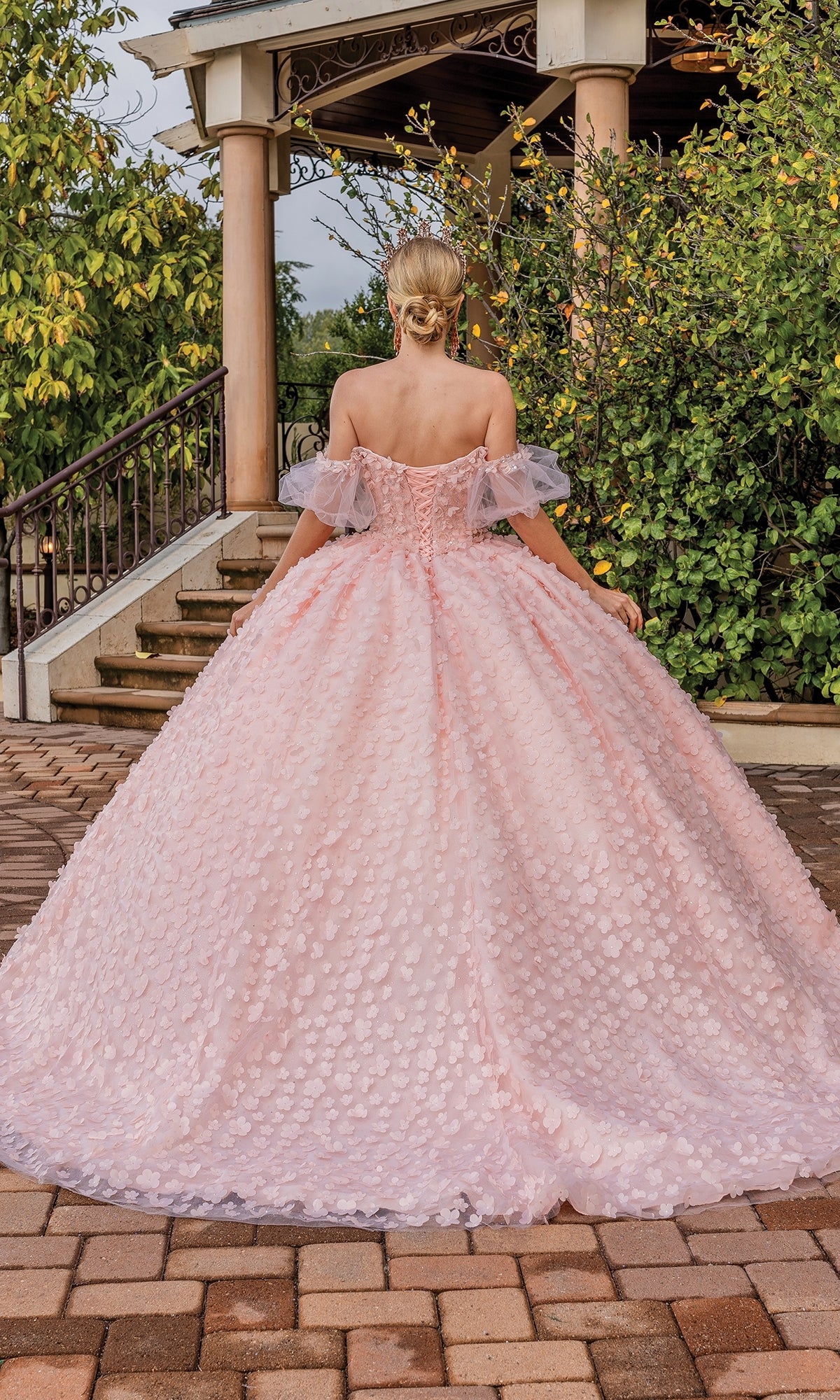 A woman wearing the Quinceanera Dress 1810 by Dancing Queen, featuring an off-the-shoulder and sweetheart neckline, stands outdoors with her back to the camera near a gazebo and greenery.