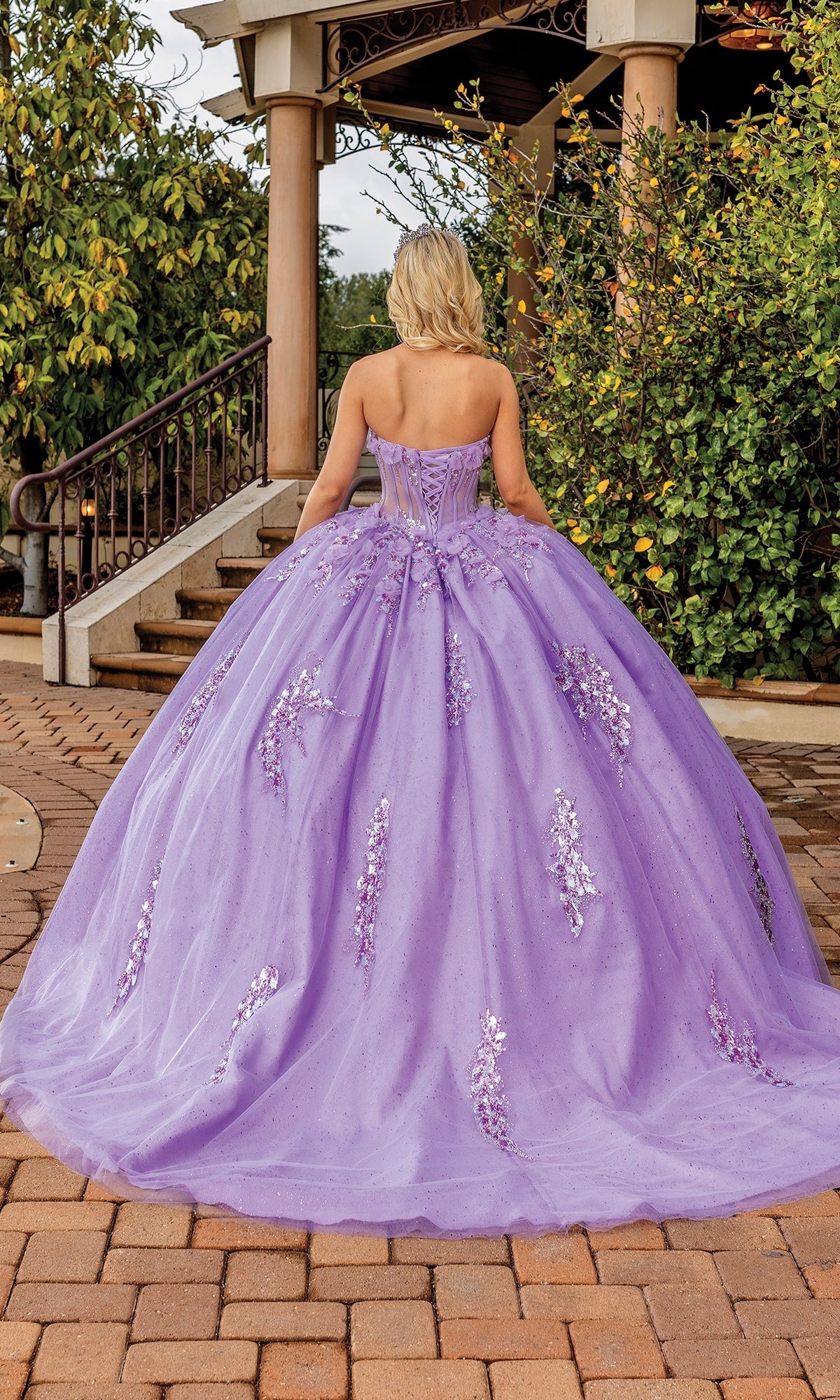 A person wearing the Quinceanera Dress 1813 By Dancing Queen, a pastel ball gown with a sequined sweetheart neckline, stands on a brick walkway facing away, with greenery and stairs in the background.