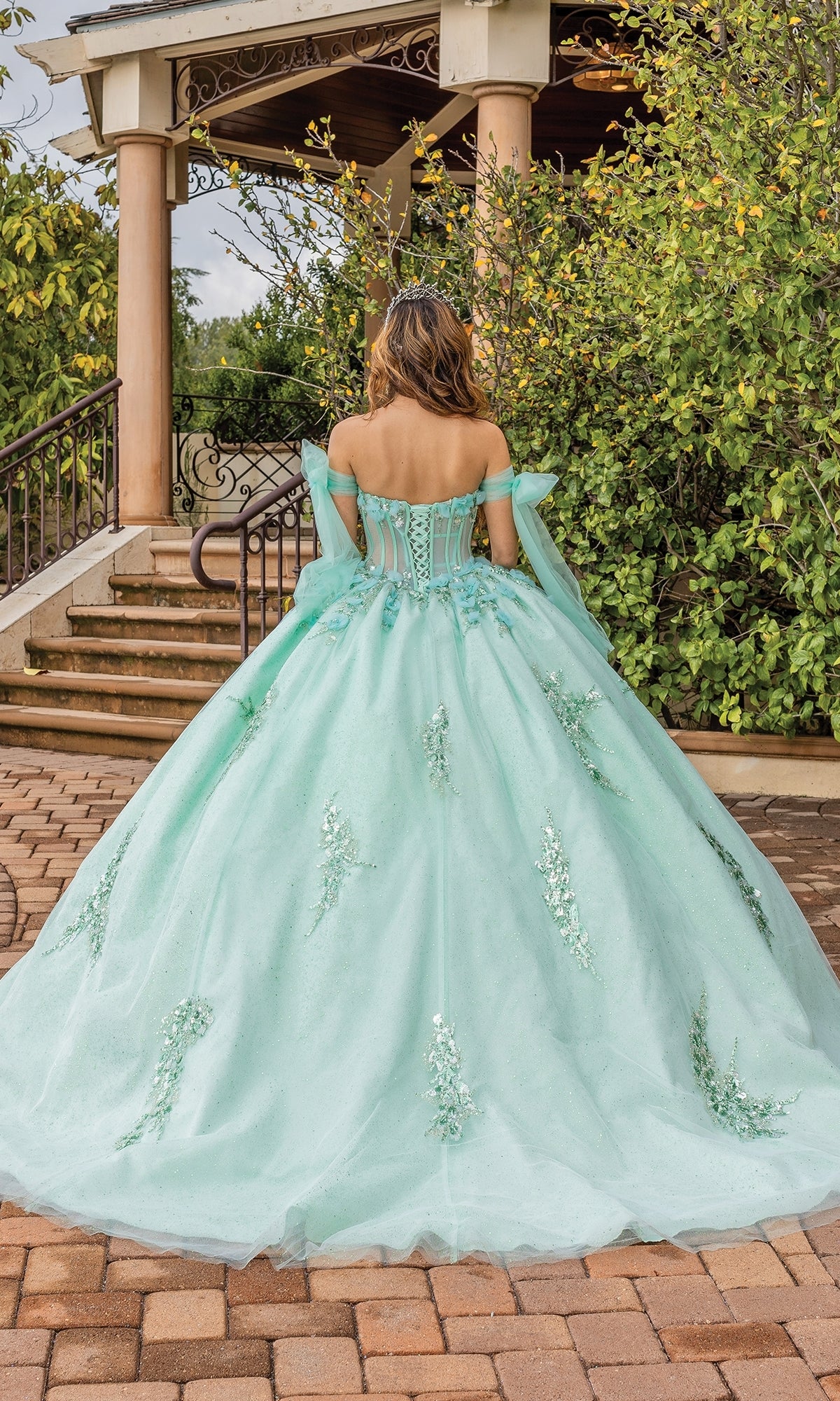 Someone wearing the Quinceanera Dress 1813 by Dancing Queen, a pastel ball gown with off-the-shoulder sleeves, stands outdoors on a stone path facing away from the camera.