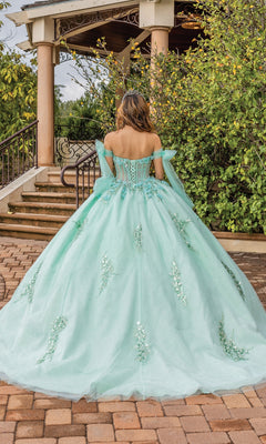 Someone wearing the Quinceanera Dress 1813 by Dancing Queen, a pastel ball gown with off-the-shoulder sleeves, stands outdoors on a stone path facing away from the camera.
