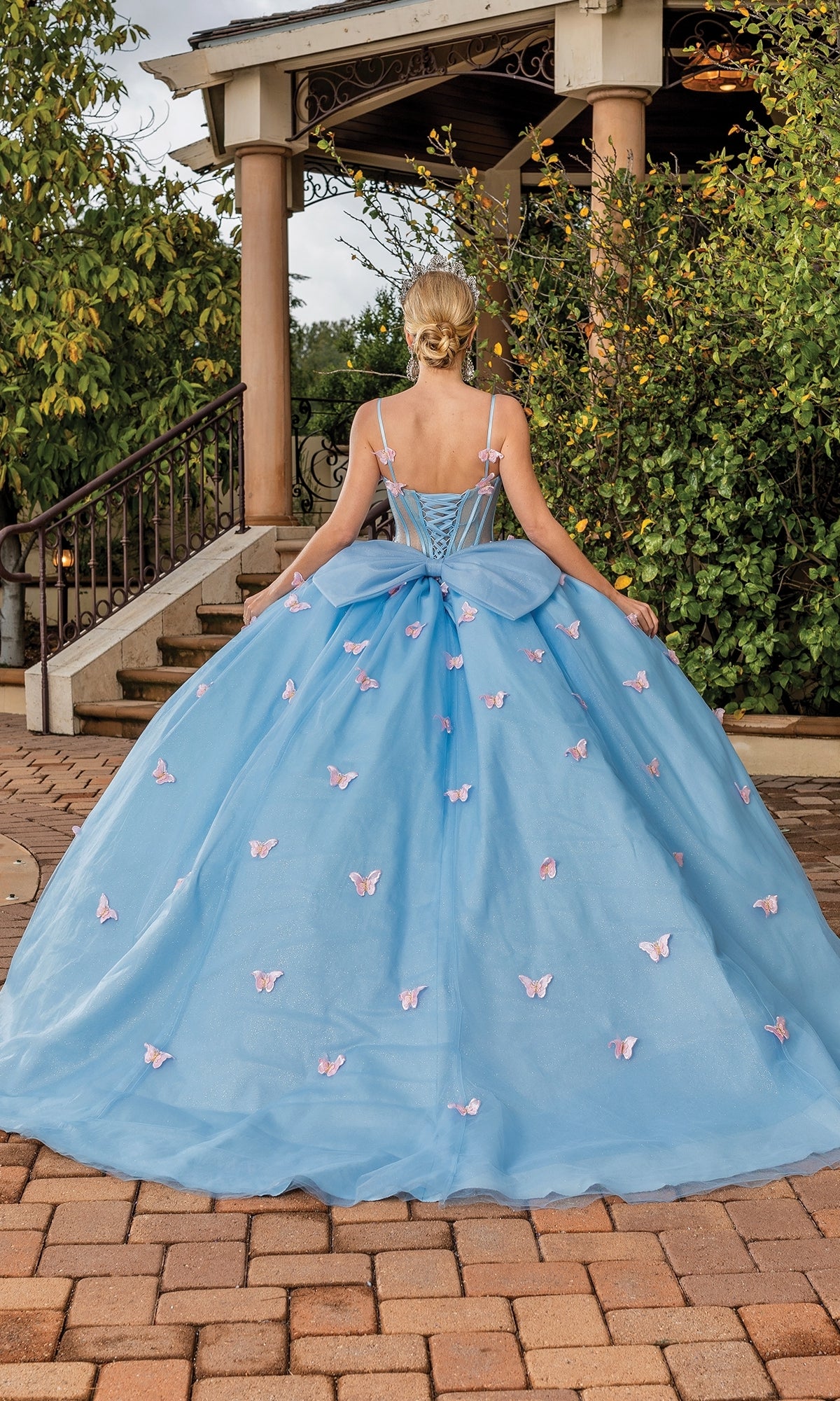 A woman with blonde hair in an updo stands outdoors with her back to the camera, wearing Quinceanera Dress 1815 By Dancing Queen—a blue ball gown decorated with pink butterflies and a dramatic bow back.