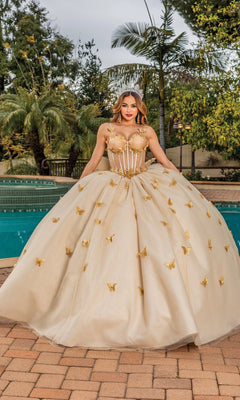A woman stands by a pool in the Quinceanera Dress 1815 By Dancing Queen, featuring gold fabric, butterfly embellishments, a sheer corset bodice, and sweetheart neckline. Palm trees and lush greenery frame the elegant scene.