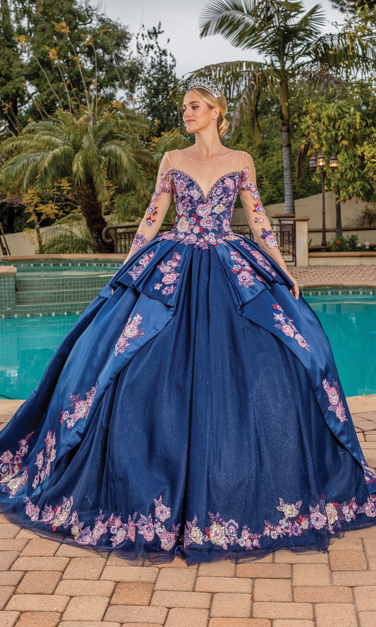 A woman in the Quinceanera Dress 1816 By Dancing Queen, featuring floral embellished sleeves and a tiara, stands by a pool with palm trees in the background.