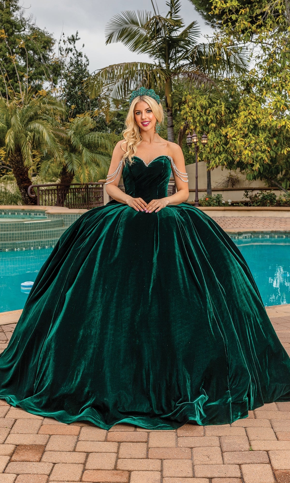 Wearing the Quinceanera Dress 1817 by Dancing Queen— a dark green velvet gown with a strapless sweetheart neckline— a woman stands on a poolside patio amid palm trees and lush greenery.