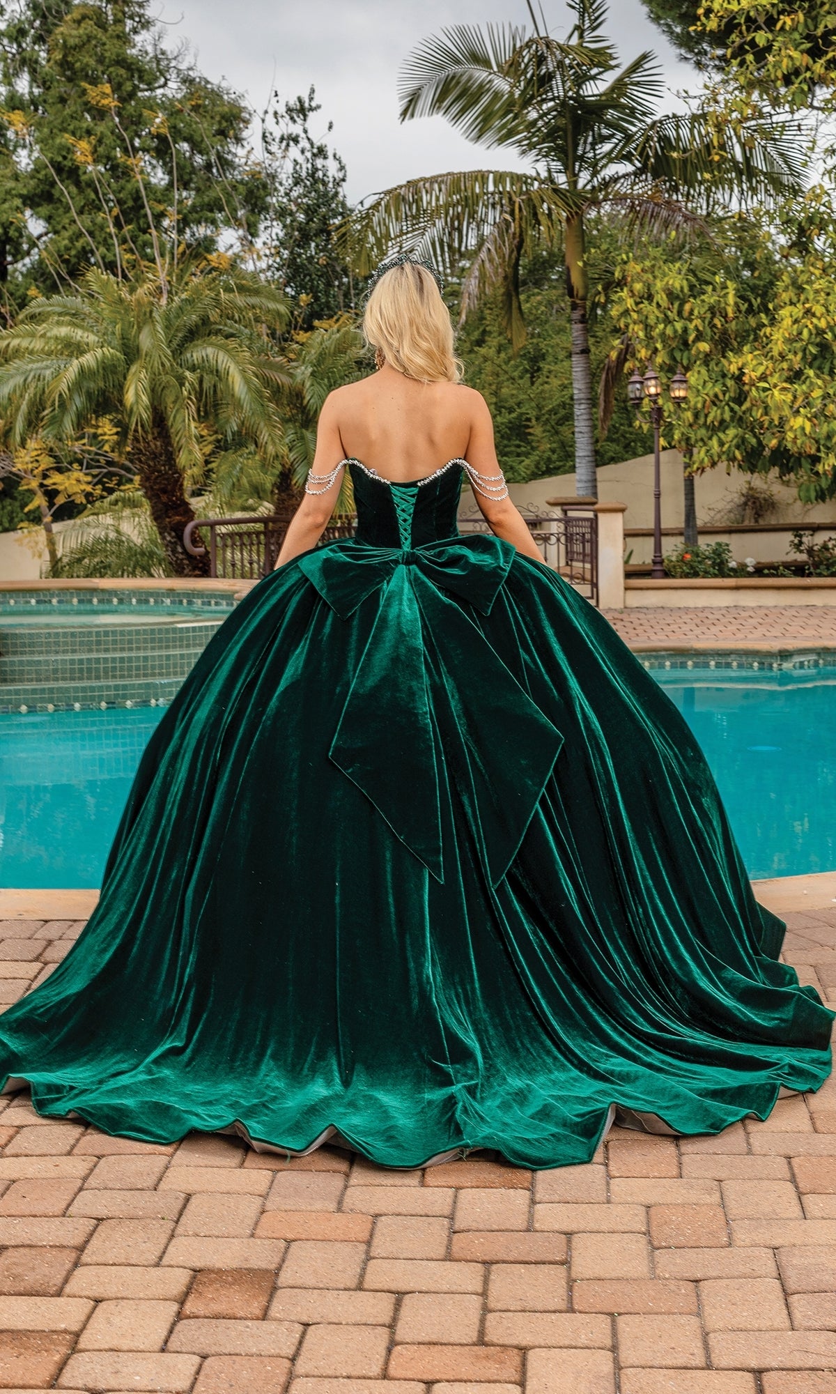Wearing the Quinceanera Dress 1817 By Dancing Queen in emerald green velvet with a strapless sweetheart neckline, a person stands by a pool facing away, with trees and a brick patio in the background.