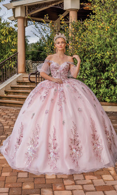A woman stands outdoors by a staircase and greenery in the Quinceanera Dress 1821 By Dancing Queen, a blush pink ball gown with a sweetheart neckline, off-the-shoulder floral embellishments, and a tiara for an enchanting look.