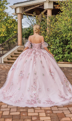 A woman wearing the Quinceanera Dress 1821 by Dancing Queen, a blush pink off-the-shoulder ball gown with floral embroidery, stands outdoors facing away before stone steps and lush greenery.