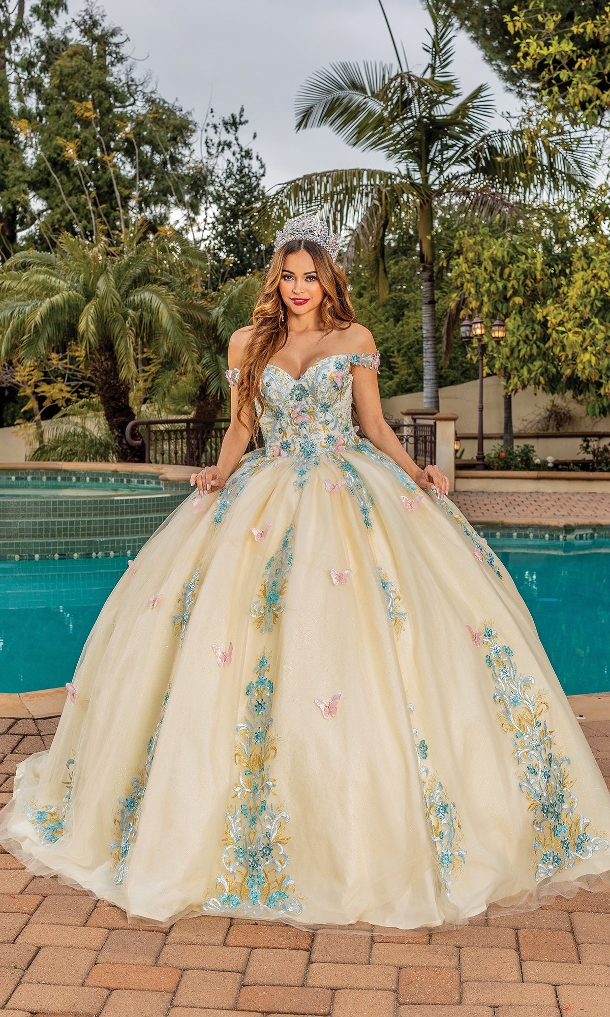 A woman stands by a pool wearing the Quinceanera Dress 1823 By Dancing Queen, a cream off-the-shoulder ball gown with blue and pink floral embroidery, framed by palm trees and lush greenery.