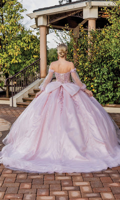 A woman wearing the Quinceanera Dress 1824 by Dancing Queen, a blush pink ball gown with off-the-shoulder straps and a large bow, stands outdoors before a gazebo, facing away from the camera.