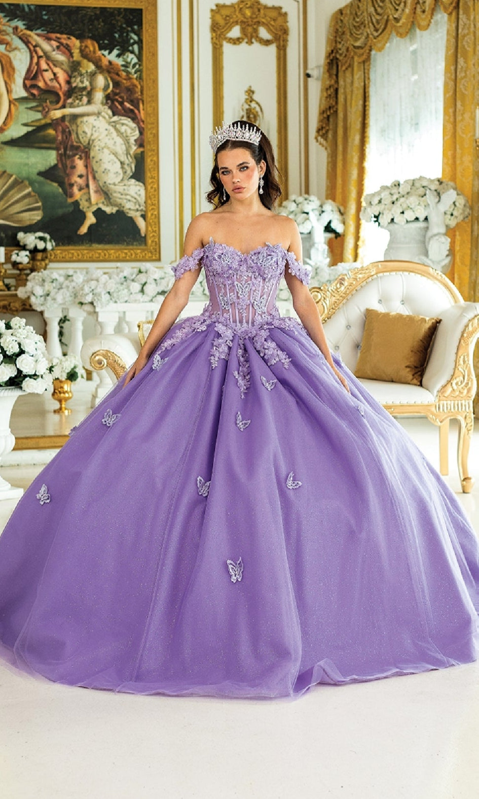 A woman wears the Quinceanera Dress 1826 By Dancing Queen, an off-the-shoulder gown with butterfly accents, standing in an ornate room with gold décor, white flowers, classical artwork, and a tiara.