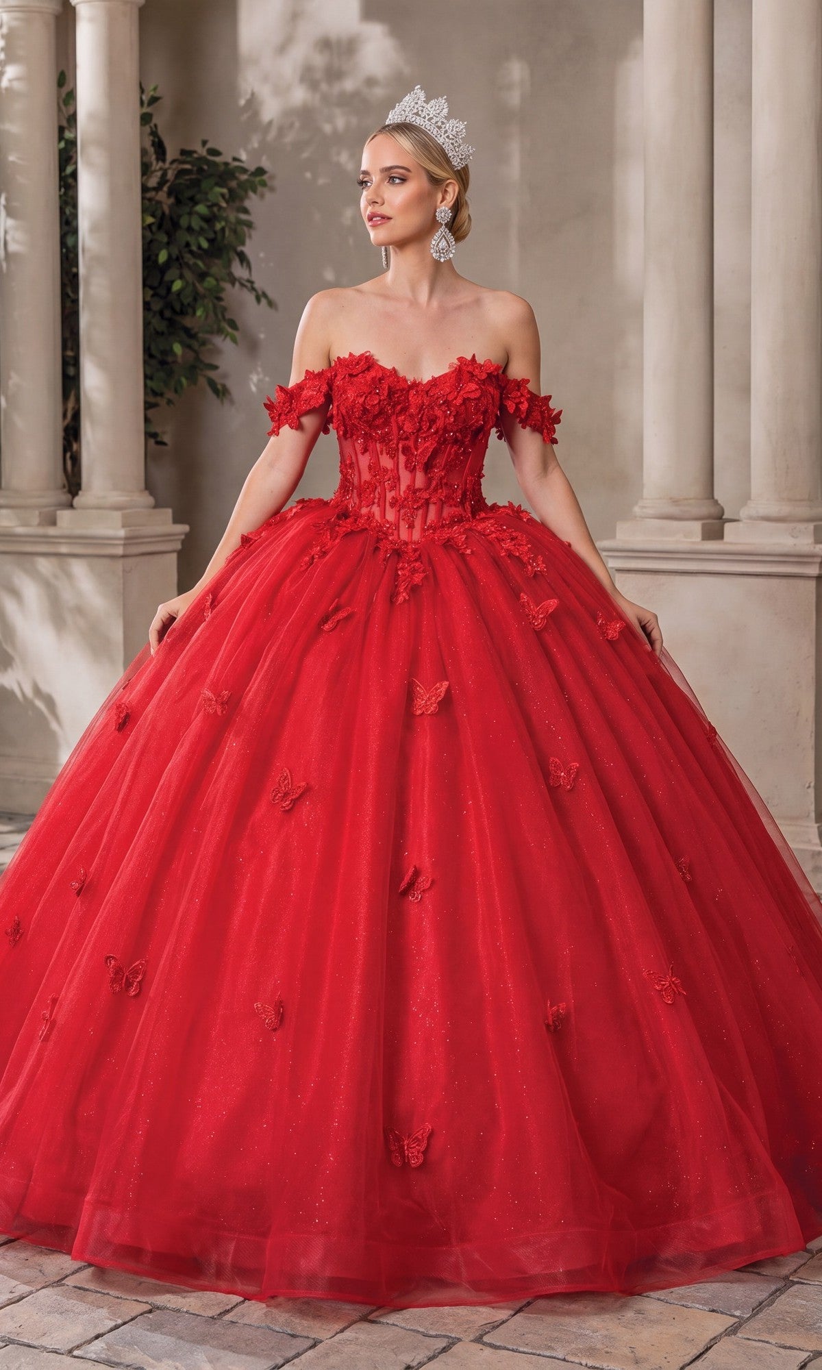 A woman stands on a stone floor surrounded by columns and lush greenery, wearing the Quinceanera Dress 1826 By Dancing Queen—a stunning red off-the-shoulder A-line ball gown adorned with floral accents and delicate lace applique—her look completed by a sparkling tiara.