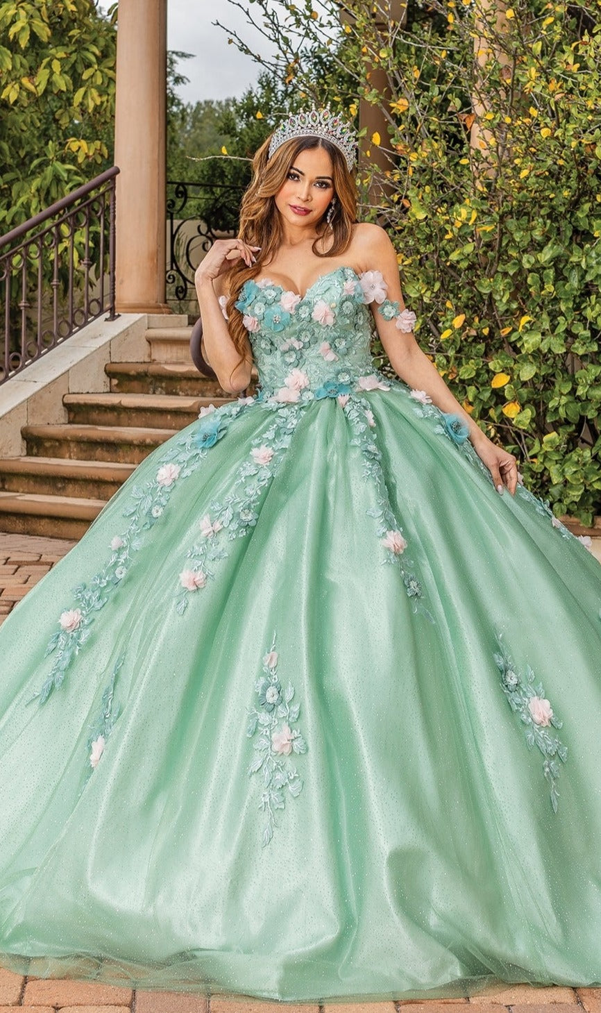 A woman in the Quinceanera Dress 1828 By Dancing Queen with floral appliqués and a tiara poses outdoors on a stone path in front of stairs and greenery.