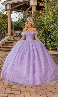 A woman stands outdoors in the Quinceanera Dress 1829 By Dancing Queen, featuring a voluminous off-the-shoulder silhouette and sweetheart neckline, with a matching tiara. She smiles with greenery and a gazebo in the background.