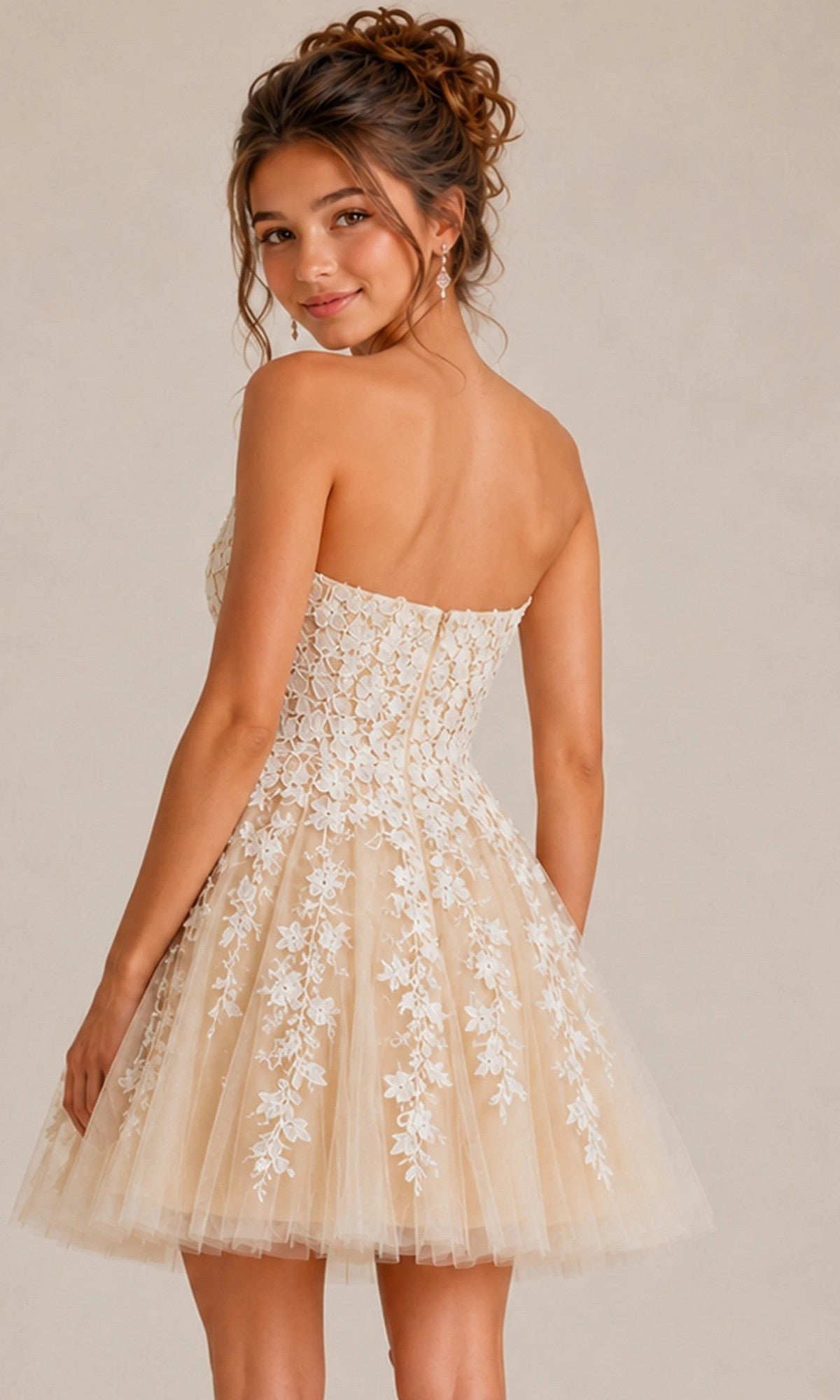 A woman is standing and looking over her shoulder against a plain background, wearing the Jovani 1830 short homecoming dress—a strapless fit-and-flare beige style adorned with white floral lace appliques.