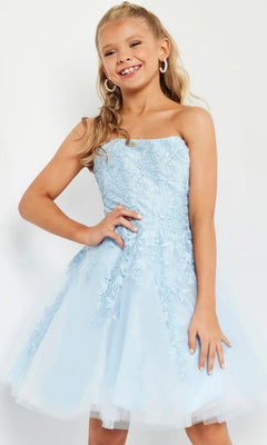 A young girl stands and smiles with one hand on her hip against a plain background, wearing the Jovani 1830 short homecoming dress: a strapless, light blue fit-and-flare design adorned with delicate floral lace appliques and layered tulle.