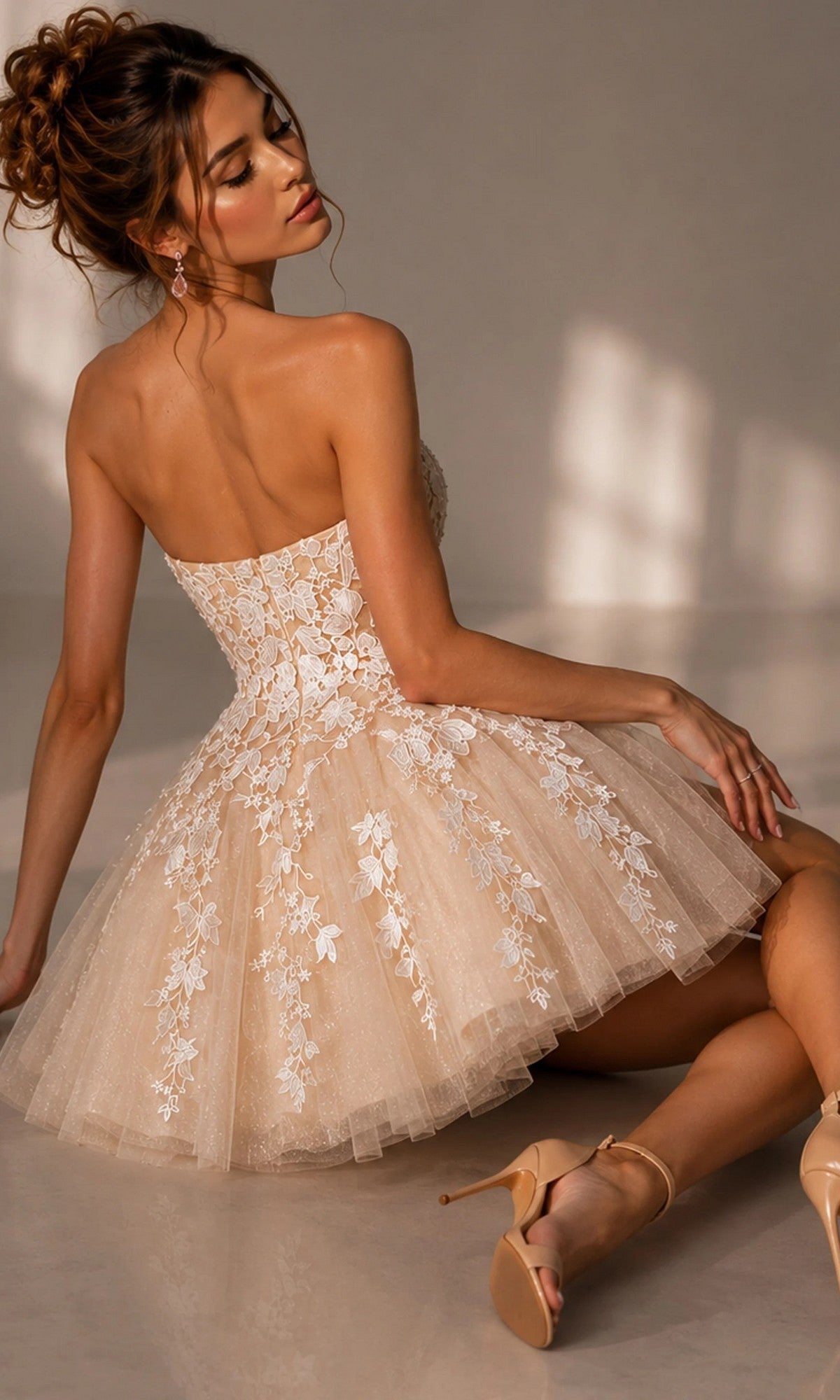 A woman sits on the floor wearing the Jovani 1830 short homecoming dress, a strapless fit-and-flare style with beige floral lace appliqués over a short tulle skirt, paired with nude high-heeled shoes.