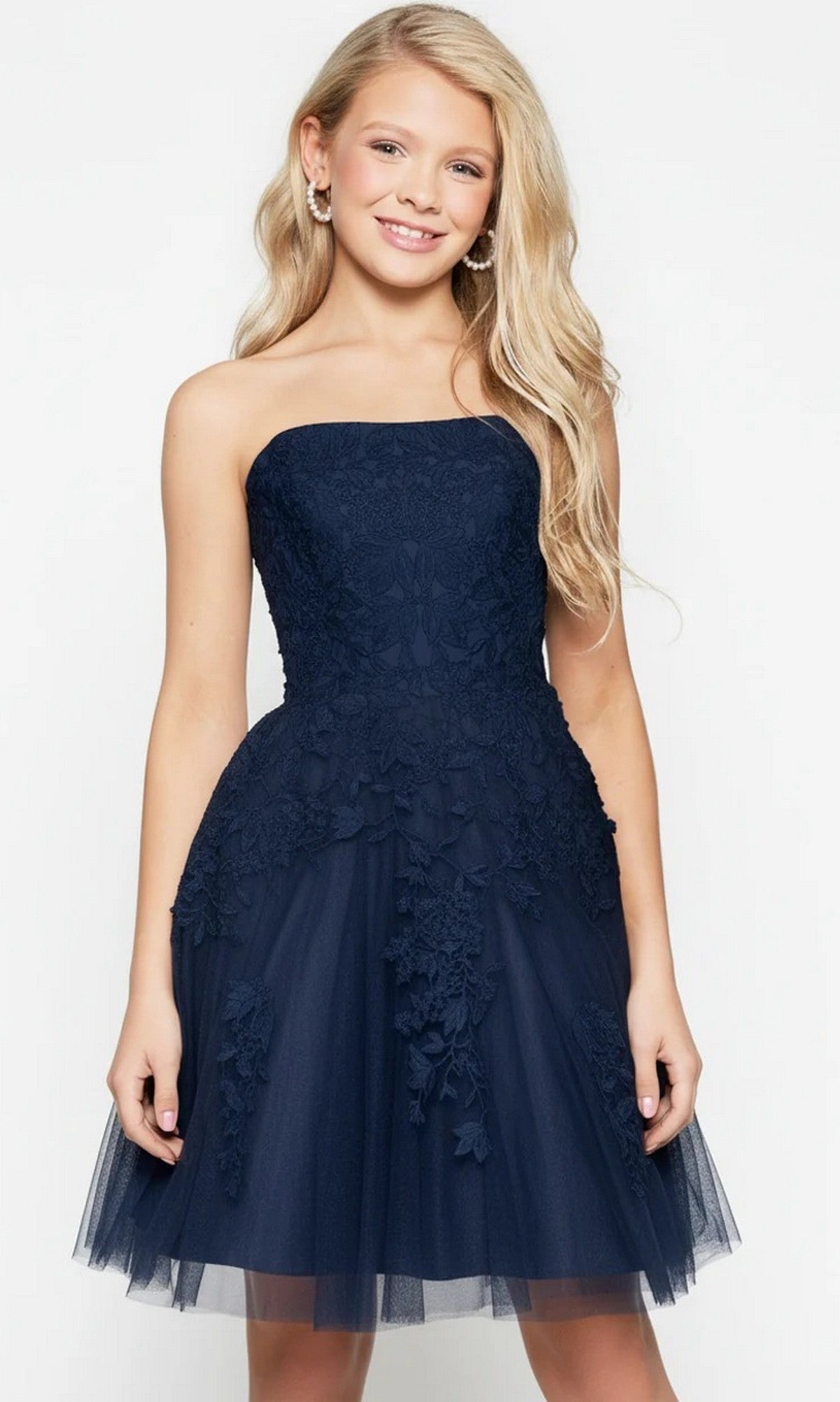 A young girl with long blonde hair stands facing forward and smiling against a plain light background, wearing the Jovani 1830 short homecoming dress, which features a strapless design adorned with delicate floral lace appliques.