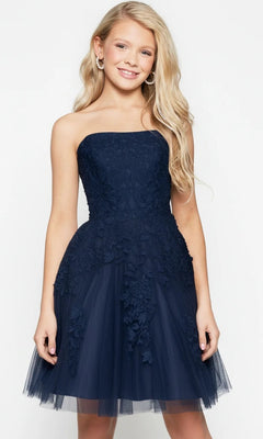 A young girl with long blonde hair stands facing forward and smiling against a plain light background, wearing the Jovani 1830 short homecoming dress, which features a strapless design adorned with delicate floral lace appliques.