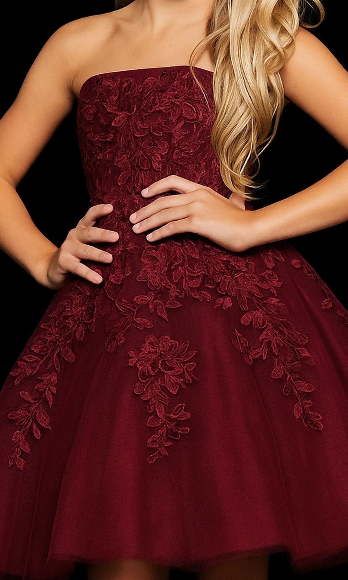 A woman poses with one hand on her hip against a black background, wearing the Jovani 1830 short homecoming dress in burgundy—a strapless fit-and-flare style adorned with intricate floral lace appliques.