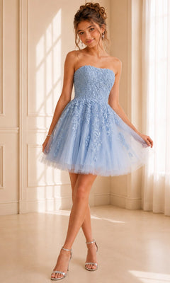 Wearing the Jovani 1830 Short Homecoming Dress, a woman showcases its strapless fit-and-flare silhouette, adorned with light blue floral lace appliques and a tulle skirt. She completes her look with silver heels while standing indoors by a sunlit window.