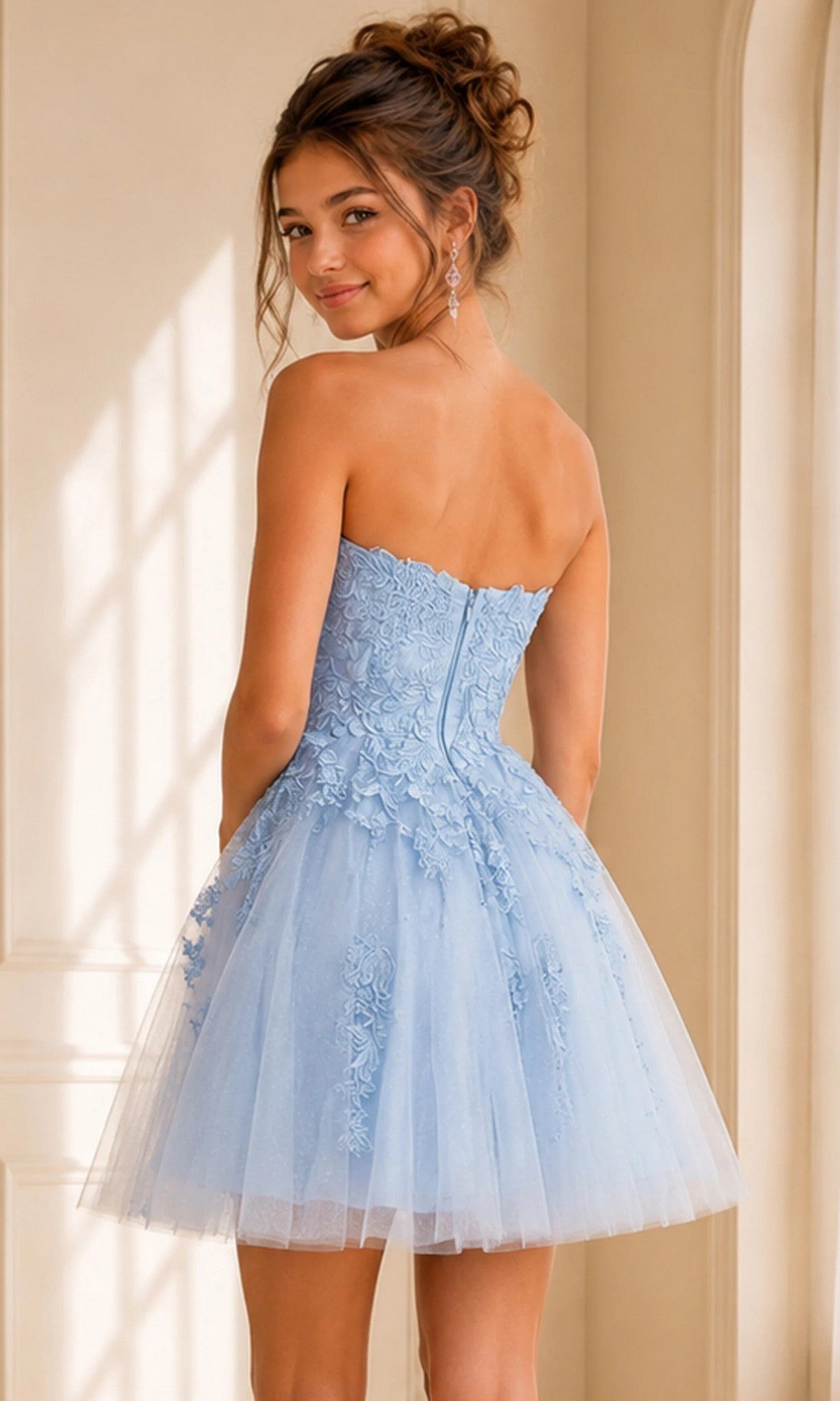 A young woman stands indoors, facing away but glancing over her shoulder, wearing the Short Homecoming Dress: Jovani 1830—a light blue, strapless fit-and-flare style adorned with intricate floral lace appliques and delicate tulle.