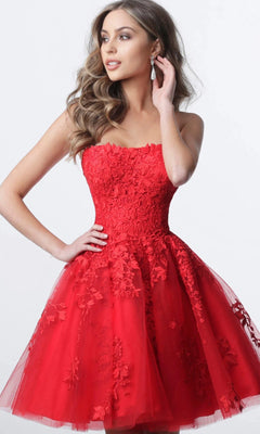 A woman models the Jovani 1830 Short Homecoming Dress in vibrant red, showcasing its strapless, fit-and-flare knee-length silhouette adorned with elegant floral lace appliques. She poses confidently with one hand on her hip and the other brushing her hair, set against a simple light background.