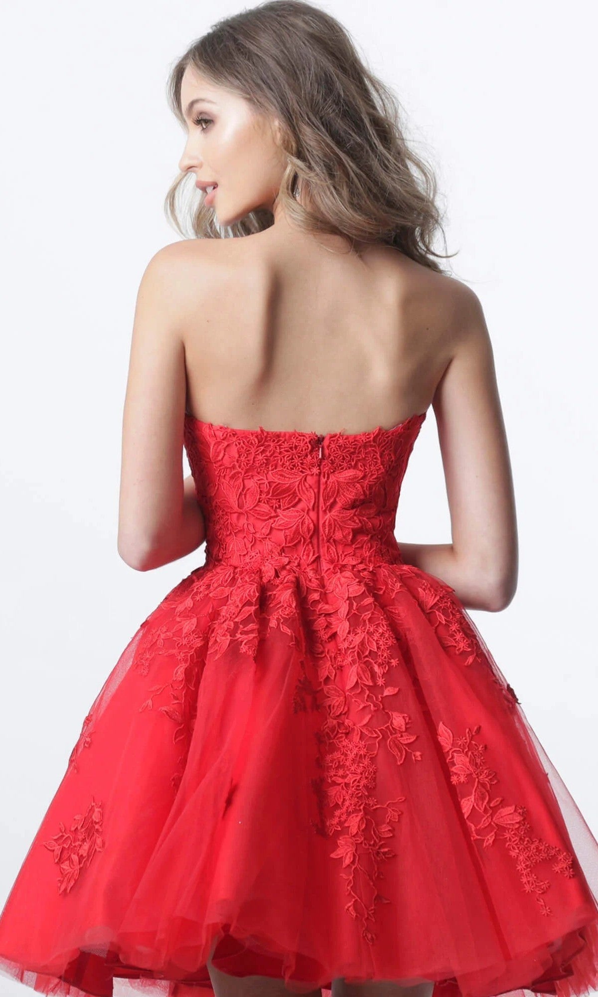 A woman wearing the Short Homecoming Dress: Jovani 1830, a strapless fit-and-flare design adorned with vibrant red floral lace appliques and embroidery, stands with her back to the camera against a plain white background.