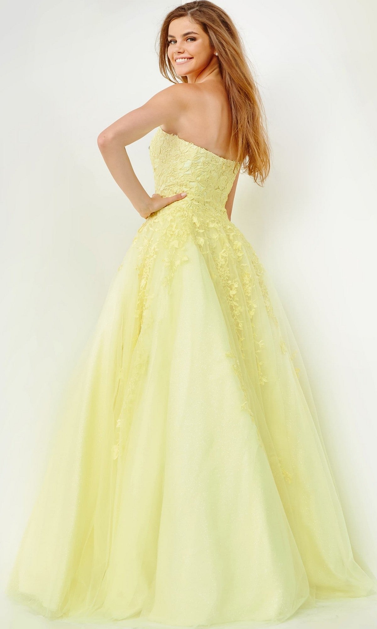 A woman wearing the Jovani 1831 long formal dress, a pale yellow, strapless, floor-length ballgown adorned with delicate floral details, stands smiling and facing away from the camera against a plain white background.