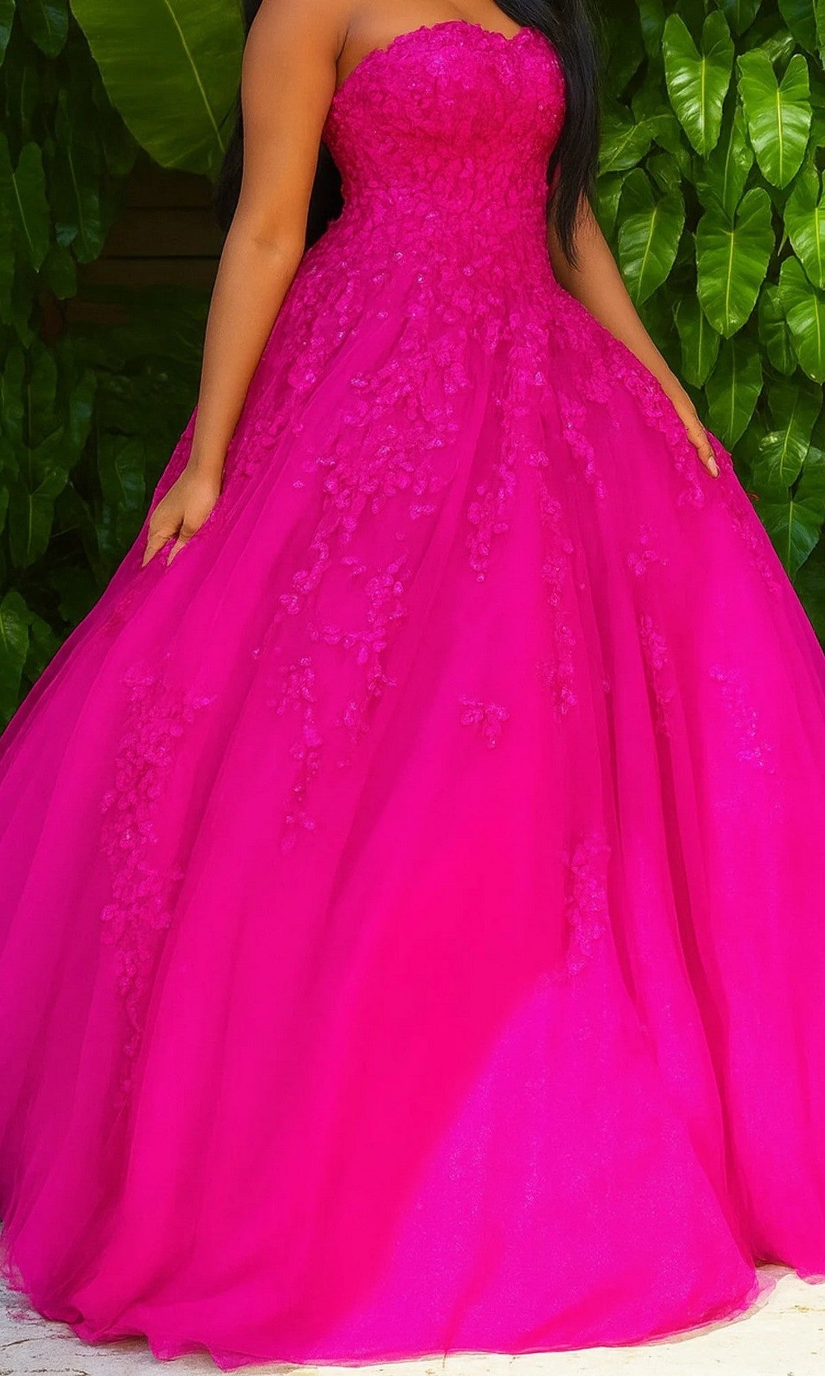 A woman wearing the Long Formal Dress: Jovani 1831 in hot pink, a strapless ballgown with textured floral details, stands in front of large green leaves.