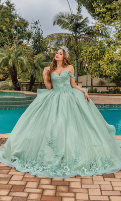 A woman in the Quinceanera Dress 1833 by Dancing Queen, a sage green ball gown with floral details and a sweetheart neckline, sits beside a pool surrounded by palm trees.