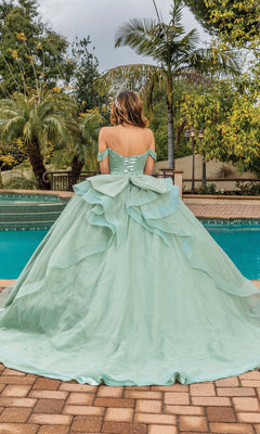 A woman in the Quinceanera Dress 1833 By Dancing Queen, a sage green ball gown with an off-the-shoulder sweetheart neckline, stands by the pool with her back to the camera, surrounded by palm trees and lush greenery.