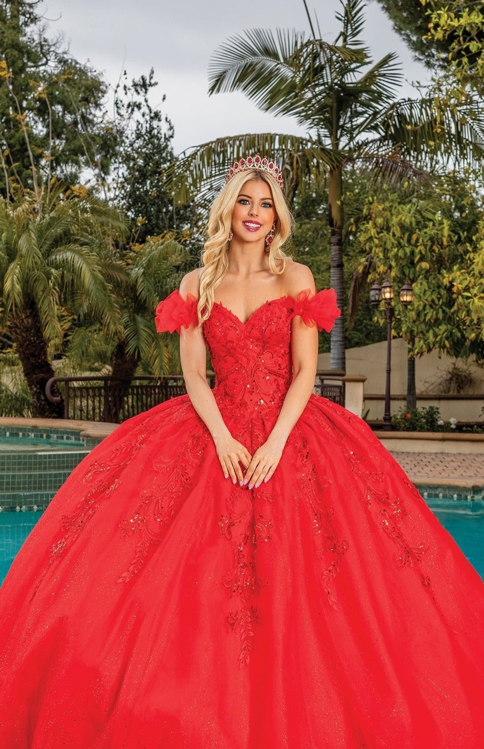 A woman wearing the Quinceanera Dress 1834 By Dancing Queen and a tiara stands in front of a pool, surrounded by palm trees and lush greenery.