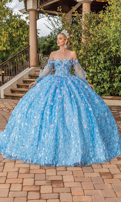 A woman stands outdoors in the Quinceanera Dress 1835 By Dancing Queen—a voluminous blue strapless sweetheart gown with floral details, removable off-the-shoulder sleeves, and a sparkling tiara for added elegance.