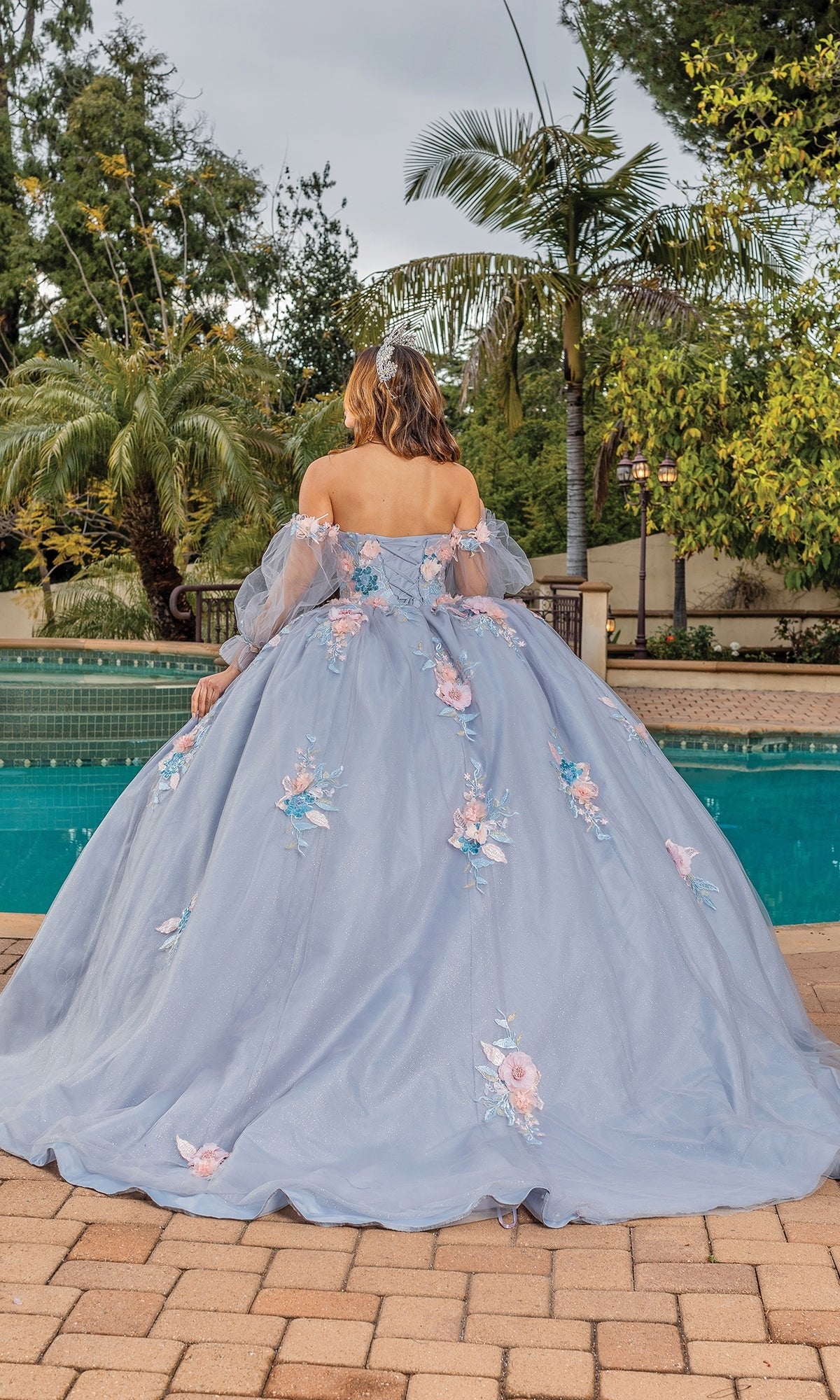 A person stands by a pool, facing away, wearing the Quinceanera Dress 1837 By Dancing Queen—a light blue ball gown with a sweetheart neckline and floral appliqués. Palm trees and lush greenery fill the background.