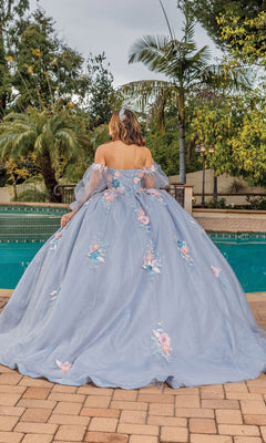 A person stands by a pool, facing away, wearing the Quinceanera Dress 1837 By Dancing Queen—a light blue ball gown with a sweetheart neckline and floral appliqués. Palm trees and lush greenery fill the background.