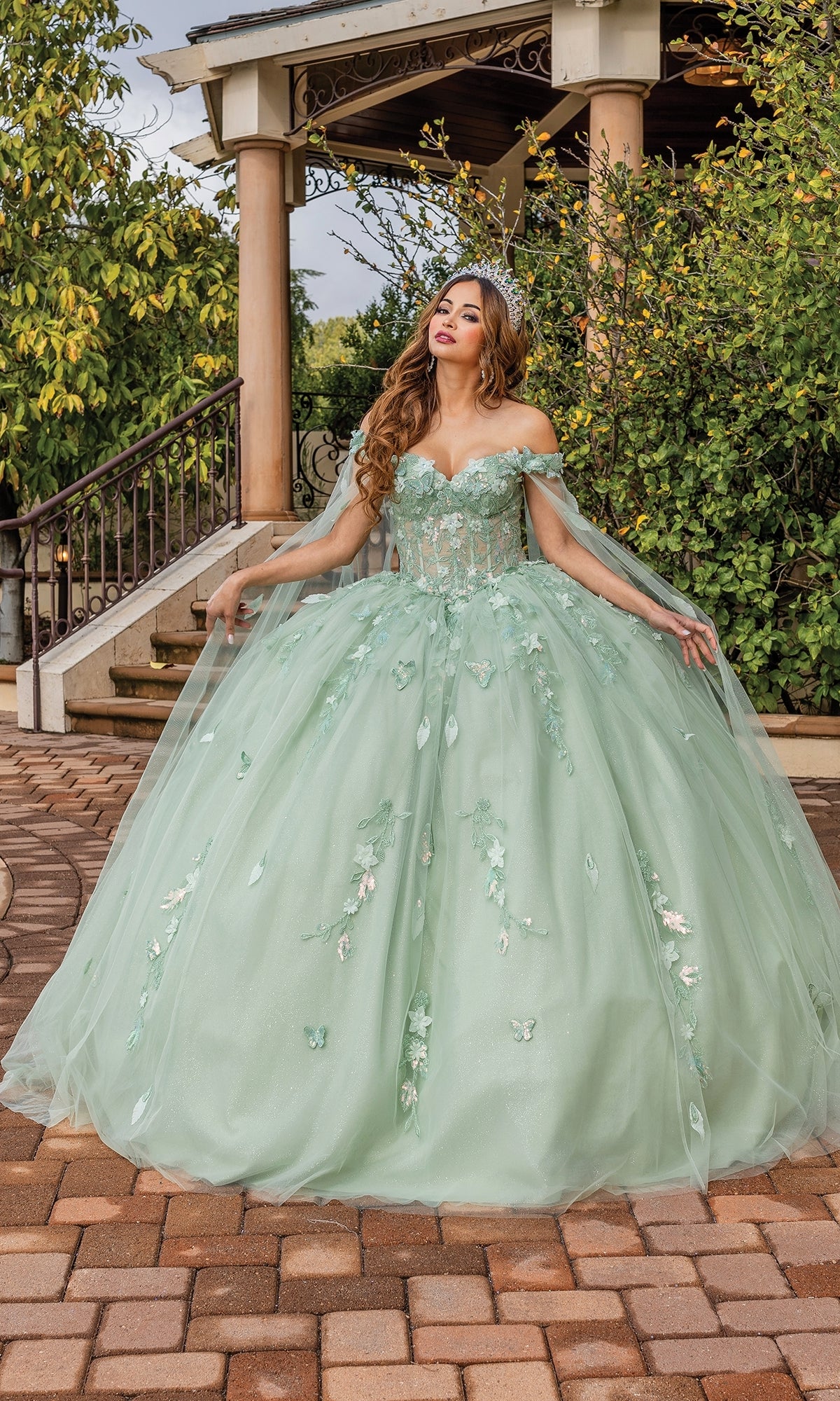 A woman stands outdoors in front of stairs, wearing the Quinceanera Dress 1840 By Dancing Queen—a light green, off-the-shoulder ball gown with floral appliqués, full tulle skirt, and a stunning sweetheart neckline.