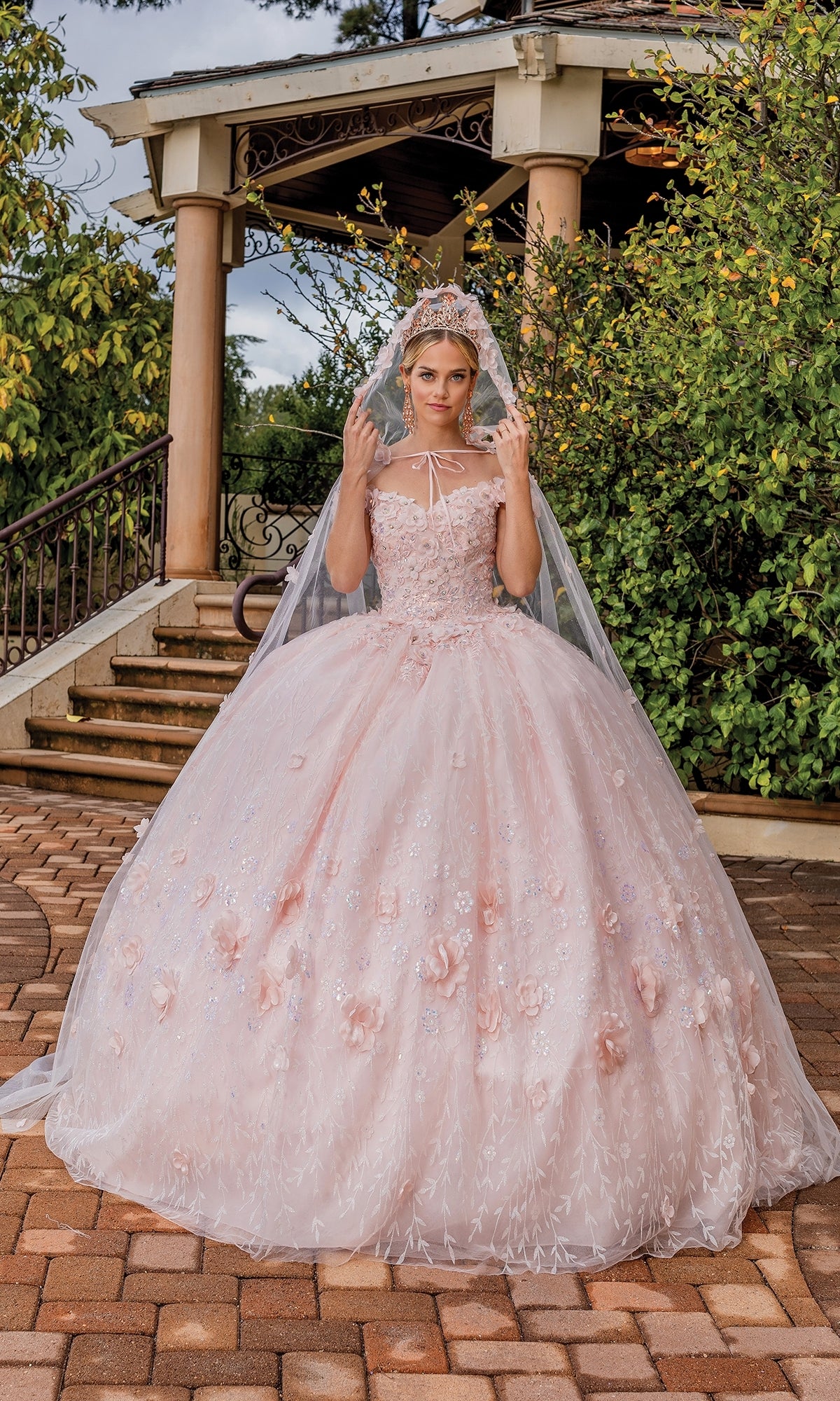 A young woman wears the Quinceanera Dress 1842 By Dancing Queen, an off-the-shoulder pink a-line ball gown with floral details, a tiara, veil, and removable cape, standing on a brick walkway near outdoor steps and a gazebo.