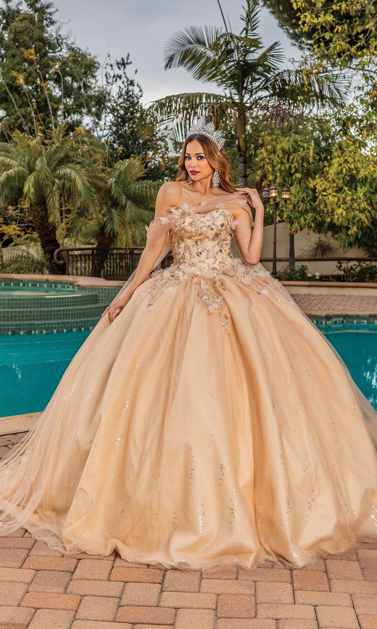 A woman poses by a pool amid palm trees and greenery, wearing the Quinceanera Dress 1844 By Dancing Queen—a peach off-shoulder ball gown with a sweetheart neckline and floral details.
