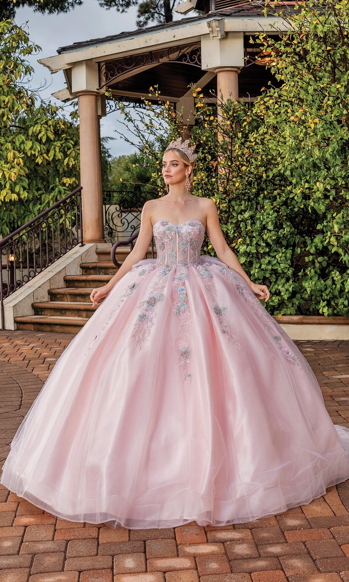 A woman stands outdoors on a brick path near a gazebo wearing the Quinceanera Dress 1848 By Dancing Queen, a strapless pale pink A-line ball gown with floral embroidery and a bow back.