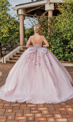 A woman faces away in front of a garden gazebo wearing the Quinceanera Dress 1848 By Dancing Queen, a light pink A-line ball gown with floral embroidery and a dramatic bow back.