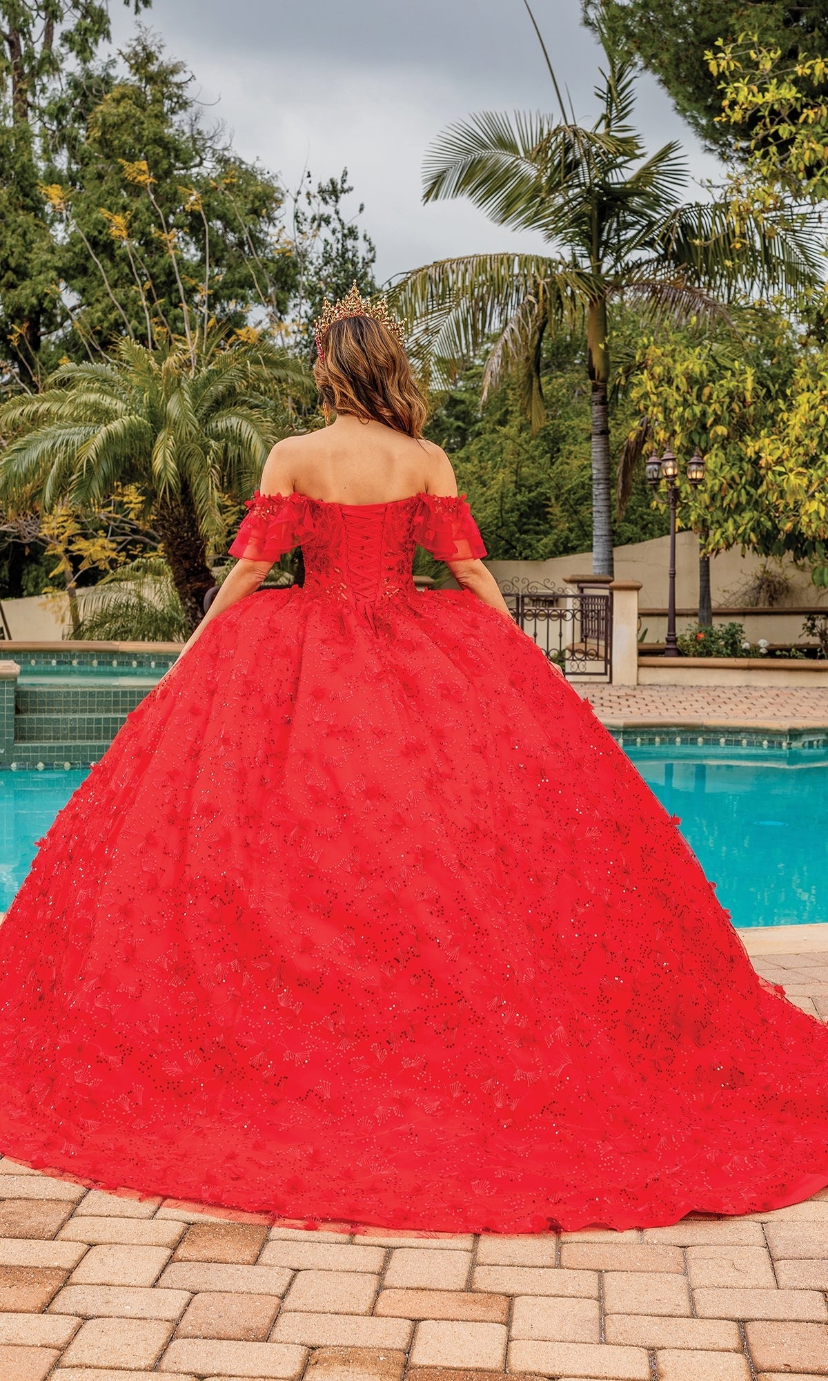 A woman in the Quinceanera Dress 1849 By Dancing Queen faces away from the camera beside a pool, surrounded by palm trees and lush greenery.